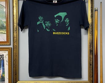 May include: A navy blue t-shirt featuring a green and black graphic of four people. The word "BUZZCOCKS" is printed in yellow below the graphic. The t-shirt is hanging on a metal bar.