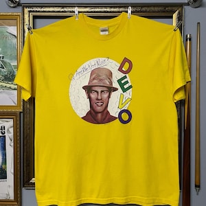May include: A bright yellow short-sleeve t-shirt featuring a graphic print of a person wearing a hat inside a white circle with golf ball dimples. The text Q: Are We Not Men? is printed above the head, and the word DEVO is printed vertically.