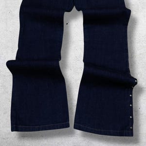 Puede incluir: Vaqueros de mezclilla azul oscuro con dise&ntilde;o de pernera acampanada. Los vaqueros tienen remaches plateados a lo largo de las costuras laterales y en los bolsillos traseros. La tela parece ser de mezclilla de peso medio.