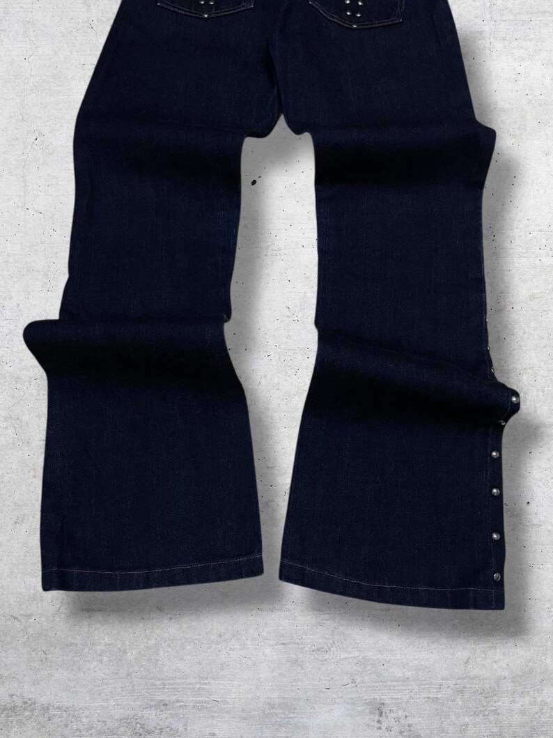 Puede incluir: Vaqueros de mezclilla azul oscuro con dise&ntilde;o de pernera acampanada. Los vaqueros tienen remaches plateados a lo largo de las costuras laterales y en los bolsillos traseros. La tela parece ser de mezclilla de peso medio.