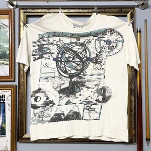 May include: Off-white t-shirt featuring a detailed map and globe design in shades of blue and gray. The shirt has a crew neck and short sleeves. The text "Gianfranco Ferre" is printed at the top.