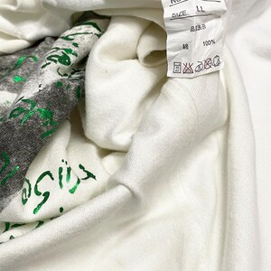 May include: A close-up of a white t-shirt with a green graphic print. The shirt has a tag with Japanese text and care symbols. The fabric is 100% cotton. The shirt is folded, showing the inside.