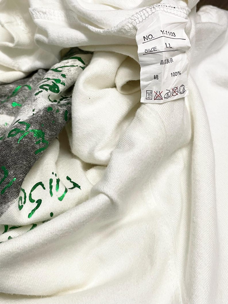 May include: A close-up of a white t-shirt with a green graphic print. The shirt has a tag with Japanese text and care symbols. The fabric is 100% cotton. The shirt is folded, showing the inside.