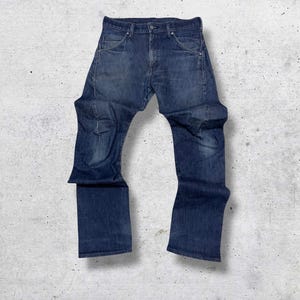 May include: Dark blue denim jeans. The jeans feature a classic five-pocket design with a button and zip closure. The denim has a faded appearance, with lighter areas on the thighs and knees. The jeans are laid flat on a light grey surface.