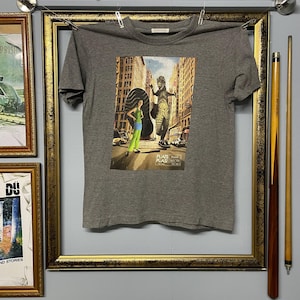 Pode incluir: Uma camiseta cinza mesclada com uma estampa gráfica colorida. O gráfico mostra uma pessoa com roupas verdes e azuis em uma rua da cidade com um monstro grande. O texto "PLATS PLASI" está impresso no gráfico.