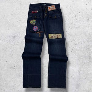 Puede incluir: Vaqueros de mezclilla azul oscuro con múltiples bolsillos y parches decorativos. Los parches incluyen un corazón, un signo de la paz y una etiqueta. Los vaqueros tienen bragueta con botones y están doblados en la parte inferior. Estilo vintage.