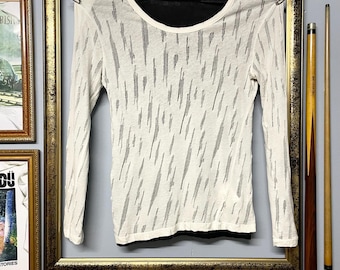 May include: A white long-sleeved shirt with a black underlayer and a crew neck. The white fabric features a pattern of vertical black lines. The shirt is hanging on a metal rod inside a gold-framed picture.