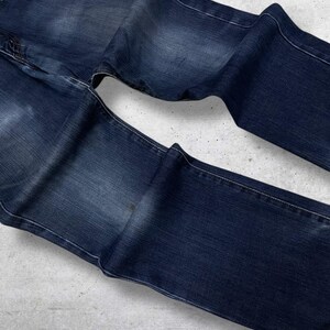 May include: Dark blue denim jeans with white stitching. The jeans are folded and laid flat, showcasing the front and back pockets and the overall shape. The denim has a slightly faded appearance.