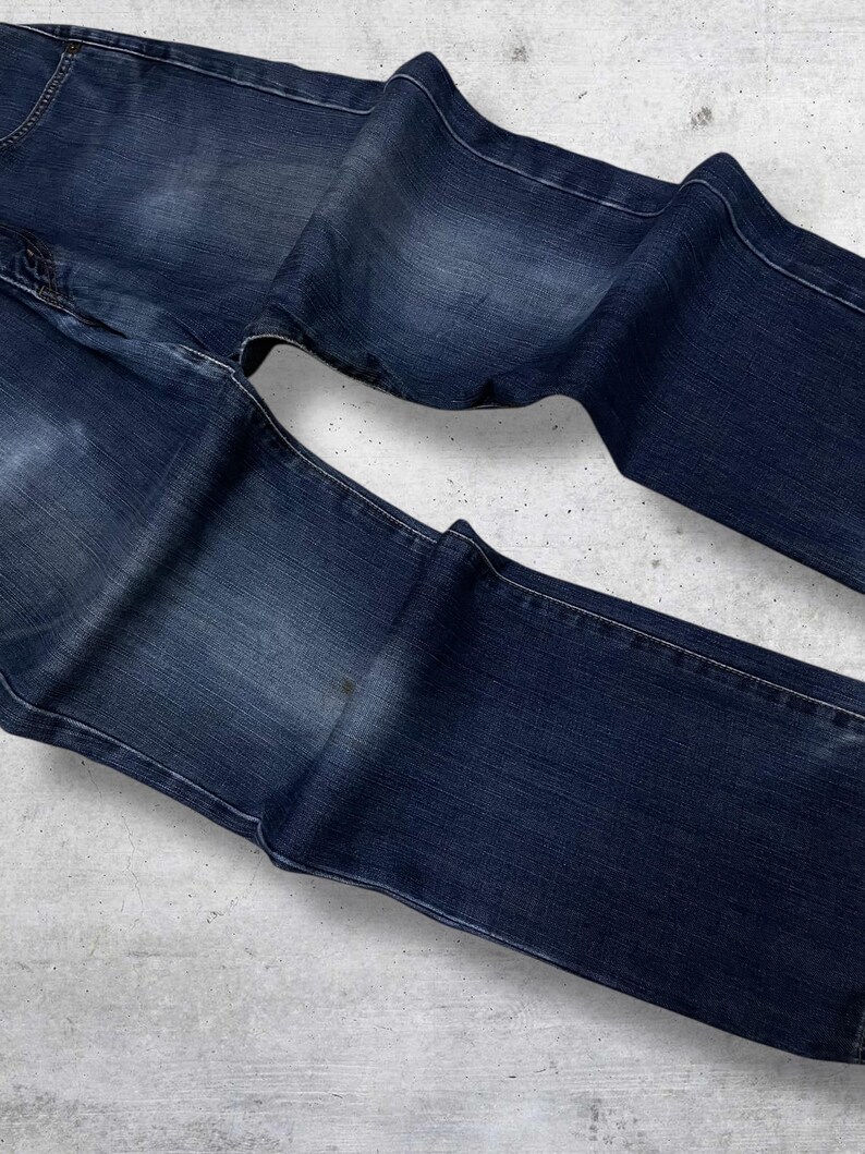 May include: Dark blue denim jeans with white stitching. The jeans are folded and laid flat, showcasing the front and back pockets and the overall shape. The denim has a slightly faded appearance.