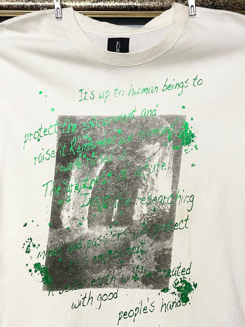 May include: Cream-coloured t-shirt featuring green handwritten text and a grayscale image. The text includes phrases like "It's up to human beings to protect the environment" and "people's hands."