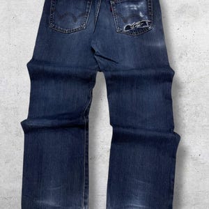May include: Dark blue denim jeans with a faded appearance and distressed details. The jeans feature a classic five-pocket design, a leather patch with the brand name "EDWIN", and a red tag on the back pocket. The jeans have a straight leg cut.