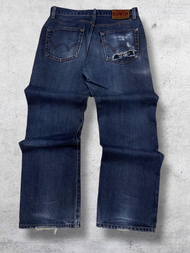 May include: Dark blue denim jeans with a faded appearance and distressed details. The jeans feature a classic five-pocket design, a leather patch with the brand name "EDWIN", and a red tag on the back pocket. The jeans have a straight leg cut.