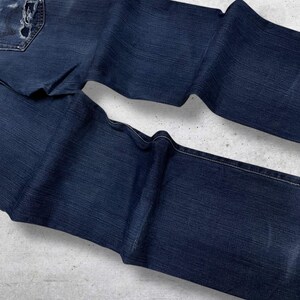 May include: Dark blue denim jeans with a distressed pocket and a faded appearance. The jeans are folded and laid out on a light gray surface, showcasing the texture and details of the fabric.