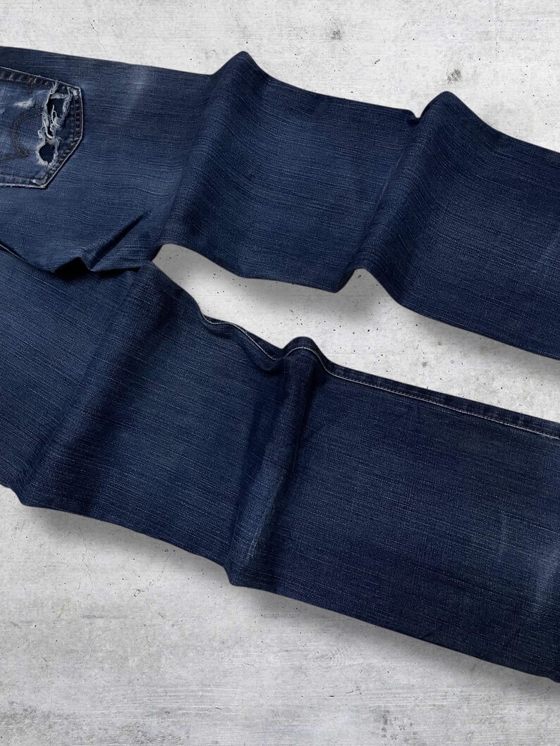 May include: Dark blue denim jeans with a distressed pocket and a faded appearance. The jeans are folded and laid out on a light gray surface, showcasing the texture and details of the fabric.