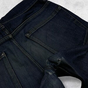 Op de afbeelding: Close-up van een donkerblauwe denim jeans. De afbeelding benadrukt de achterzakken, naden en de textuur van de stof. De jeans vertoont tekenen van slijtage, met een distressed look.