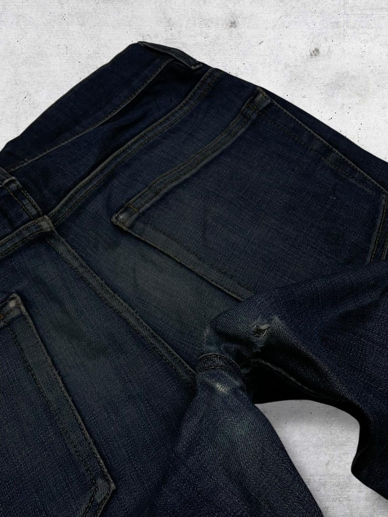 Op de afbeelding: Close-up van een donkerblauwe denim jeans. De afbeelding benadrukt de achterzakken, naden en de textuur van de stof. De jeans vertoont tekenen van slijtage, met een distressed look.