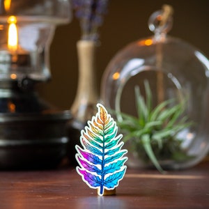 May include: A rainbow-colored fern leaf sticker on a wooden surface. The sticker is a vibrant mix of red, orange, yellow, green, blue, and purple.
