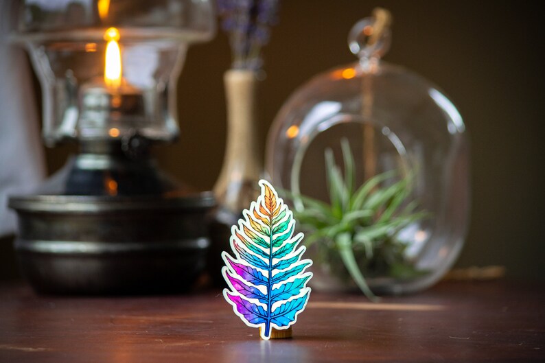 May include: A rainbow-colored fern leaf sticker on a wooden surface. The sticker is a vibrant mix of red, orange, yellow, green, blue, and purple.