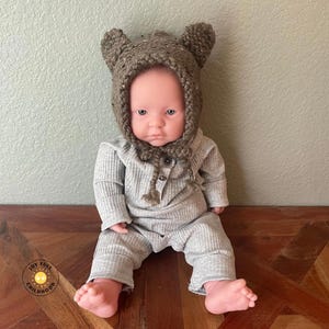 Könnte beinhalten: Eine Puppe trägt einen hellgrauen, gerippten Langarm-Strampler mit Knopfleiste und eine braune, gesprenkelte Strickmütze mit zwei kleinen, abgerundeten Ohren. Die Puppe hat einen hellen Hautton und sitzt in aufrechter Position.