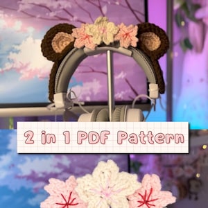 Könnte beinhalten: Zwei gehäkelte Kopfhörerbezüge. Einer ist ein braunes Bärenohren-Stirnband mit rosa und weißen Kirschblüten. Der andere ist ein rosa und weißes Kirschblüten-Stirnband. Beide sind auf einem weißen Kopfhörer zu sehen.