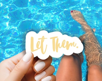 May include: A white sticker with the text "Let Them." in gold lettering. The sticker is being held by a hand with white nail polish.