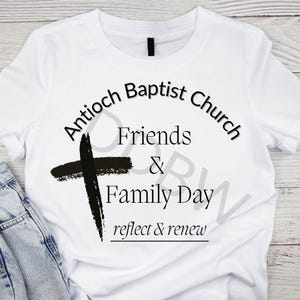 May include: White t-shirt with a black cross and text. The text reads "Antioch Baptist Church" arched at the top, with "Friends & Family Day" below, and "reflect & renew" at the bottom. The shirt is paired with blue jeans.