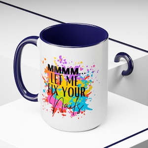 Könnte beinhalten: Weiße Keramik-Kaffeetasse mit blauem Rand und Henkel. Die Tasse hat ein buntes Farbklecks-Design und den Text "MMMM... LET ME FIX YOUR NAILS" in Schwarz.