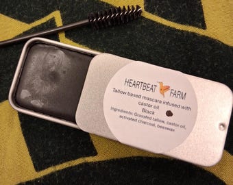 May include: A black mascara in a small, rectangular, silver tin with a matching applicator brush. The tin has a white label with the text "HEARTBEAT FARM" and product information. The mascara is tallow-based and infused with castor oil.