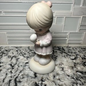 May include: A porcelain figurine of a young girl in a pink dress holding a white object. The figurine has a light-colored hairstyle and a small pink bow. It stands on a white circular base.