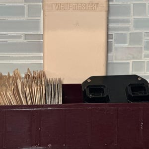 May include: A vintage View-Master set with a maroon storage box, a beige case, and a black viewer. The box contains numerous slide reels. The beige case is labeled "VIEW-MASTER".
