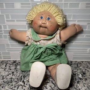May include: A vintage doll with a round face, blue eyes, and blonde yarn hair. The doll wears a white and green floral dress with a green checkered skirt. The doll is sitting with its arms outstretched and white shoes.