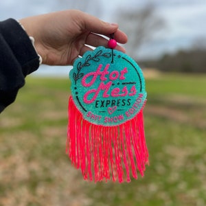 Puede incluir: Un adorno redondo turquesa con el texto "Hot Mess Express Shit Show Edition" en letras negras y rosas. El adorno tiene una franja rosa alrededor del borde.