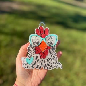 May include: A white, glittery chicken charm with black spots, wearing teal blue glasses and a red comb. The chicken has a small teal blue heart on its chest.