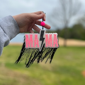 Könnte beinhalten: Ein pinkfarbenes und schwarzes Schild mit der Aufschrift "MAMA" und einem Blitz in der Mitte. Das Schild hat schwarzen Fransen an den Rändern und eine rosa Perle, die oben hängt.