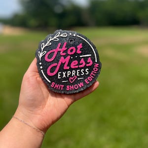 Puede incluir: Un ambientador redondo negro con el texto "Hot Mess Express Shit Show Edition" en rosa y blanco. El ambientador se sostiene en una mano sobre un fondo verde borroso.