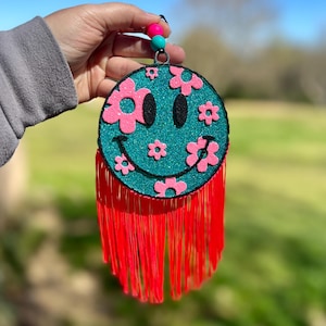 Puede incluir: Una cara sonriente azul turquesa con flores rosas y purpurina. La cara sonriente está unida a una franja roja.