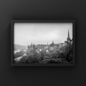 Può includere: Una fotografia in bianco e nero di uno skyline cittadino con un campanile di chiesa sullo sfondo. La città è avvolta dalla nebbia e gli edifici sono per lo più nascosti dagli alberi.