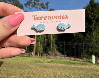 May include: A pair of light blue whale-shaped stud earrings on a pink card with the word "Terracotta" in orange. The card also has the text "HANDCRAFTED GEMS". The earrings are displayed against a background of green grass and trees.