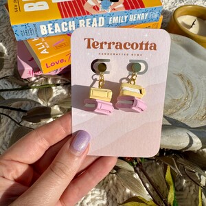 May include: A pair of pink and yellow book-shaped earrings with gold chains. The earrings are on a white card with the text "Terracotta Handcrafted Gems".