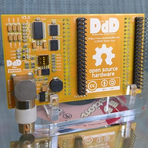May include: A yellow circuit board labeled "The Domesday Duplicator" with various electronic components. The board features the text "open source hardware" and a gear logo. It is mounted on a clear acrylic stand.