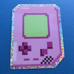 May include: A pink sticker featuring a pixelated handheld gaming device. The sticker has a green screen, purple buttons, and a silver glitter border. The design is reminiscent of retro video game consoles.