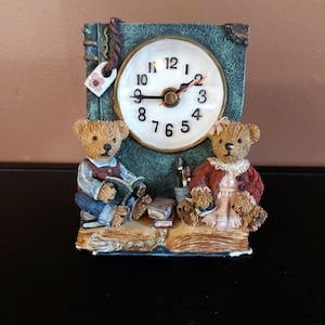 Puede incluir: Un reloj verde con forma de libro con dos osos de peluche sentados encima. La esfera del reloj tiene agujas negras y números romanos. Los osos de peluche llevan ropa y sostienen libros.