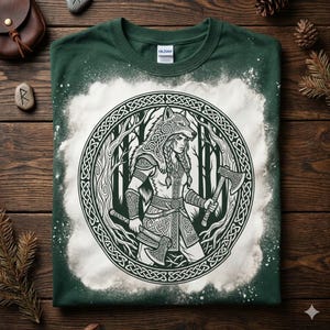 May include: Green t-shirt with a white graphic of a warrior with a wolf headdress, holding two axes, surrounded by a circular Celtic knot design. The design is set against a forest backdrop. The shirt has a bleached, distressed effect.