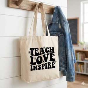 Può includere: Una borsa tote beige con le parole "TEACH LOVE INSPIRE" in nero, con un font retrò, decorata con piccoli disegni di margherite. La borsa è appesa a un gancio di legno, accanto a una giacca di jeans.