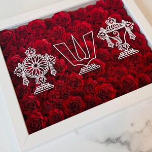 May include: A framed artwork featuring a dense arrangement of red paper roses. White cut-out designs of religious symbols are layered over the roses. The frame is white, creating a contrast with the vibrant red floral background.