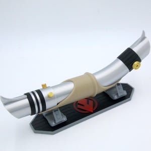 May include: A silver and black lightsaber prop with gold detailing, displayed on a black stand with a red emblem. The prop has a curved design and is made of various materials.