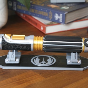 Peut inclure: Un sabre laser noir et argent avec des accents dorés est présenté sur un support noir avec le symbole Jedi. Le sabre laser a une poignée noire texturée et une lame argentée.