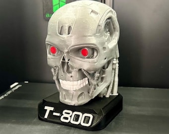 May include: A silver-colored T-800 Terminator skull model with red eyes, mounted on a black base. The base is labeled "T-800" in white text. The model is set against a dark background, possibly inside a 3D printer.