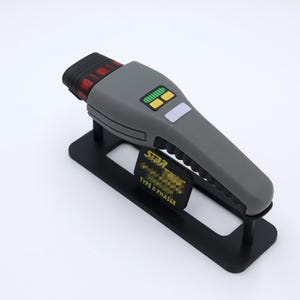 K&ouml;nnte beinhalten: Eine graue Star Trek Type II Phaser-Replik auf einem schwarzen Display-St&auml;nder. Der Phaser hat ein schwarzes und rotes Detail oben und gelbe Kn&ouml;pfe.