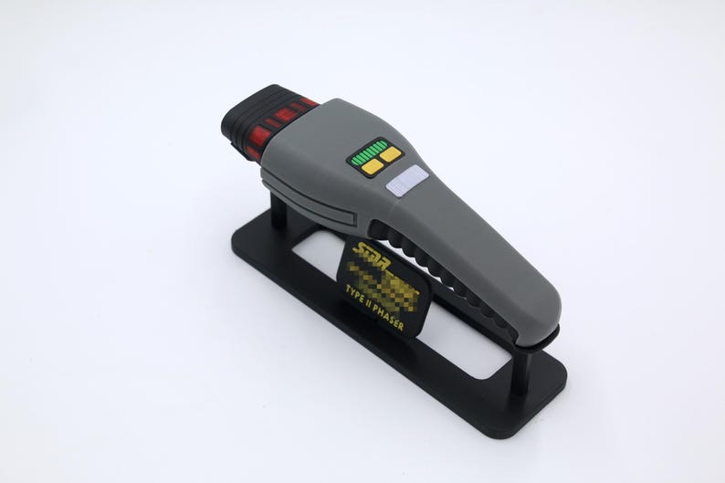 K&ouml;nnte beinhalten: Eine graue Star Trek Type II Phaser-Replik auf einem schwarzen Display-St&auml;nder. Der Phaser hat ein schwarzes und rotes Detail oben und gelbe Kn&ouml;pfe.
